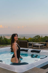 Woman in a black swimsuit standing in an outdoor hot tub on a hillside patio, gazing at a pastel sunset over rolling countryside with wooden lounge chairs in the background.