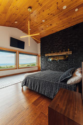 Cozy modern bedroom with vaulted pine ceiling and starburst chandelier, black stacked-stone accent wall, textured gray bedding, warm hardwood floors and panoramic windows framing rolling hills and a lake view.