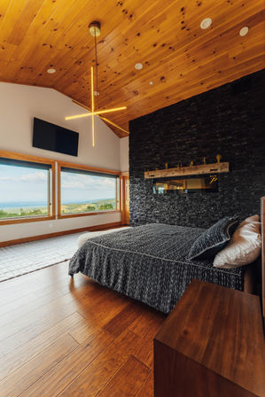 Cozy modern bedroom with vaulted pine ceiling and starburst chandelier, black stacked-stone accent wall, textured gray bedding, warm hardwood floors and panoramic windows framing rolling hills and a lake view.