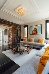 Sunlit modern living room with L-shaped gray sectional, stacked-stone accent wall, geometric glass-and-wood coffee table, crystal chandelier, beach-themed wall art and yellow accent pillows.