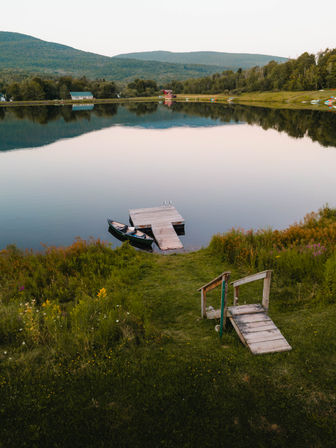 Serene mountain lake at dusk with a small wooden dock and canoe tied at the shore, grassy path and wildflowers in the foreground, tree-covered hills reflected in the calm water.