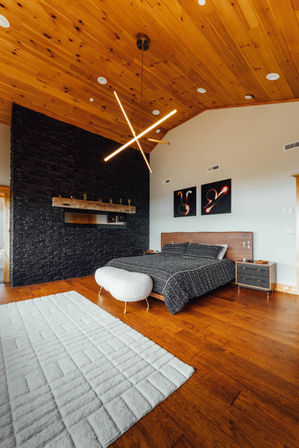 Modern master bedroom with vaulted pine ceiling, black stacked-stone accent wall, sleek crossed LED chandelier, wooden headboard with dark patterned bedding, hardwood floors and white textured area rug.
