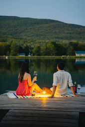 Romantic evening on a wooden dock by a calm mountain lake: two people sit on a striped blanket with wine glasses, a glowing lantern, a bottle of wine and a camera, enjoying the dusk view.