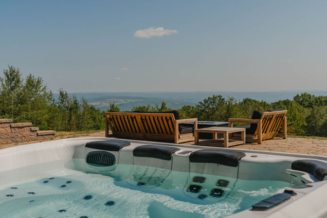 Inviting outdoor hot tub and wooden lounge seating on a stone patio overlooking rolling hills and a distant mountain valley under a clear blue sky