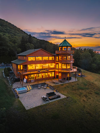 Sunset view of a glowing multi-level wooden mountain lodge with a green-roofed turret, wraparound decks, patio seating and hot tub on a grassy hillside surrounded by forest.