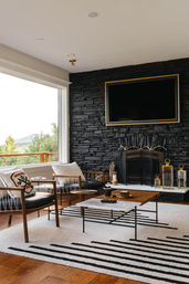 Cozy modern living room with large picture window framing a mountain view, black stacked-stone fireplace wall with wall-mounted TV, mid-century wooden lounge chairs, marble-top coffee table and striped area rug.