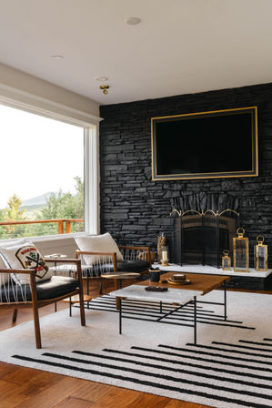 Cozy modern living room with large picture window framing a mountain view, black stacked-stone fireplace wall with wall-mounted TV, mid-century wooden lounge chairs, marble-top coffee table and striped area rug.