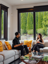 Two people relaxing with drinks on a light-gray sectional in a modern living room with large black-framed windows and lush garden view, yellow champagne-themed throw pillows and a whiskey bottle on a glass coffee table.