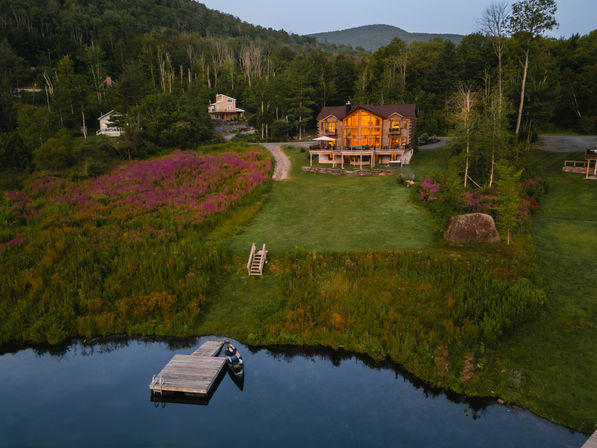 Aerial view of a lit wooden lakeside lodge with a dock and rowboat, grassy lawn, pink wildflower meadow and forested hills at dusk