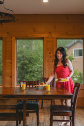 Cozy rustic cabin breakfast scene: woman in a red sundress sets plates of eggs and muffins and two glasses of orange juice on a wooden table by large windows with green trees outside.