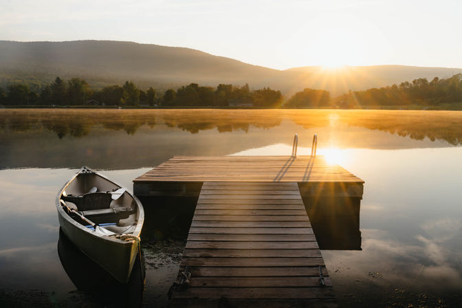 Tranquil sunrise over a misty mountain lake with a wooden dock and a canoe tied alongside, golden sunlight reflecting on calm water