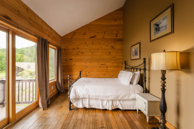 Cozy sunlit rustic cabin bedroom with wooden plank walls and floors, iron-framed single bed in white linens, bedside table and lamp, and large sliding glass doors opening to a balcony with green forest view.