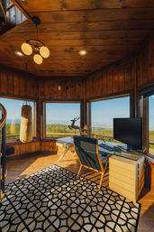 Sunlit wood-paneled home office with glass desk and monitor, leather chair, hanging punching bag, geometric rug and deer sculpture, panoramic windows overlooking rolling green hills