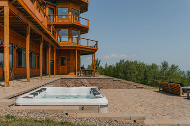 Rustic wooden lodge patio with a built-in hot tub, lounge chairs and dining table overlooking a forested hillside under a clear blue sky — perfect outdoor vacation rental scene.