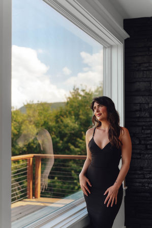 Woman in a fitted black dress standing by a large picture window in a modern home, gazing thoughtfully at sunlit trees, blue sky and a wooden deck with her reflection in the glass.