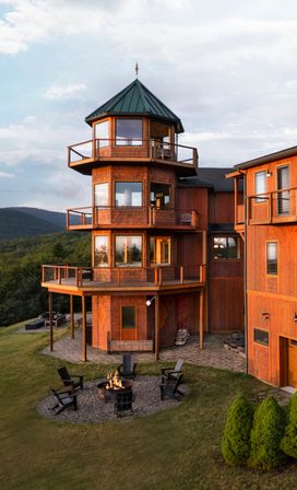 Three-story wooden turret house with green metal roof and wraparound balconies overlooking forested rolling hills, grassy yard with circular fire pit and Adirondack chairs — rustic mountain retreat