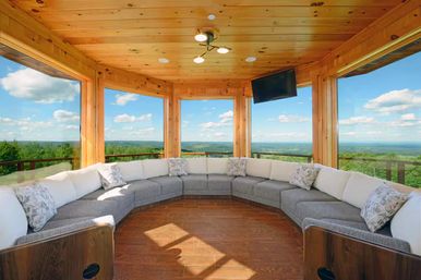 Cozy wood-paneled sunroom with panoramic windows overlooking rolling green hills and blue sky, featuring a curved gray-and-white sectional sofa, hardwood floor, and wall-mounted TV.