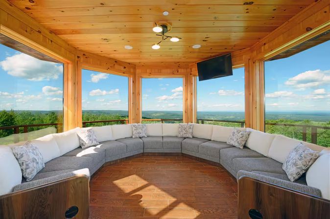 Cozy wood-paneled sunroom with panoramic windows overlooking rolling green hills and blue sky, featuring a curved gray-and-white sectional sofa, hardwood floor, and wall-mounted TV.