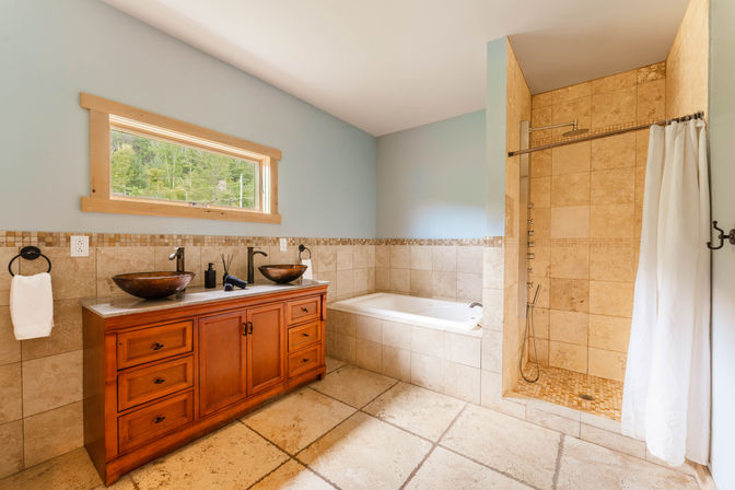 Bright spa-style bathroom with wooden double vanity and vessel sinks, beige tile floors and walls, built-in soaking tub, walk-in tiled shower with curtain, and a horizontal window showing a leafy green view.