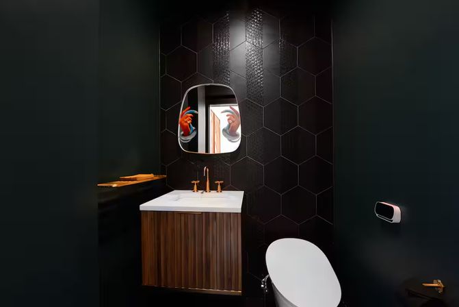 Moody modern powder room with matte black hexagon tile wall, floating wood vanity with white countertop and brass faucet, rounded mirror reflecting colorful hands, white toilet and wall-mounted dispenser — contemporary bathroom interior design