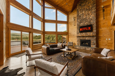 Cozy rustic lakeside cabin living room with floor-to-ceiling windows, leather sofas and shearling armchairs, patterned rug, stone fireplace with wall-mounted TV, and a wooden deck overlooking a lake and forested hills.