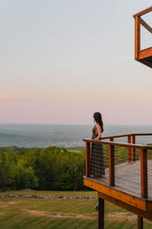Woman standing on a wooden deck, leaning on the railing and overlooking rolling green hills and distant mountains under a pastel sunset sky — scenic countryside overlook.