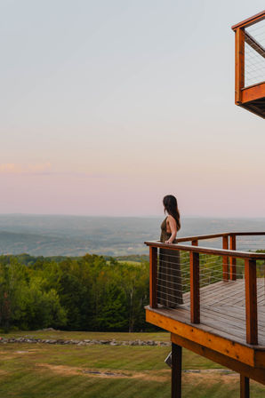 Woman standing on a wooden deck, leaning on the railing and overlooking rolling green hills and distant mountains under a pastel sunset sky — scenic countryside overlook.