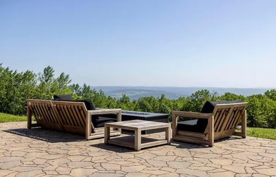 Stone hilltop patio with wooden outdoor sofas and a low coffee table around a rectangular fire pit, black cushions facing a green treeline and distant rolling hills under a clear blue sky