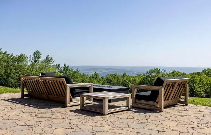 Stone hilltop patio with wooden outdoor sofas and a low coffee table around a rectangular fire pit, black cushions facing a green treeline and distant rolling hills under a clear blue sky