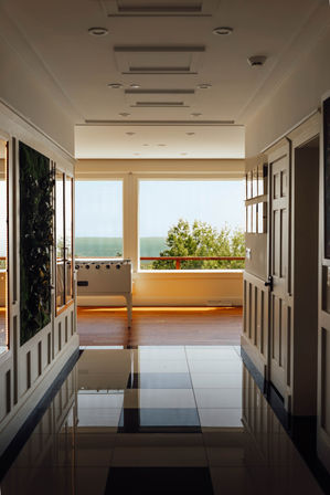 Sunlit modern hallway with glossy checkered floor leading to a coastal sitting area with large windows, a foosball table, balcony railing and ocean and treetop views.