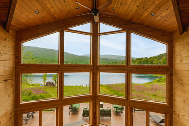 Cozy lakefront wood cabin interior with vaulted pine ceiling and expansive floor-to-ceiling windows framing a panoramic lake, rolling green hills, and a deck with lounge chairs.