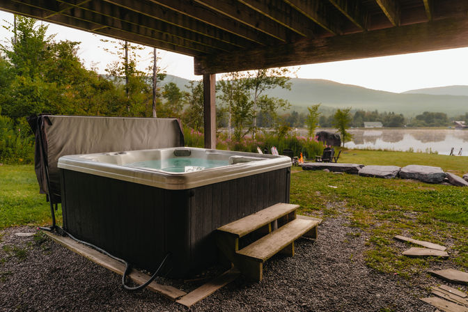 Cozy outdoor hot tub beneath a wooden deck with steps, overlooking a misty mountain lake, grassy shore, fire pit and Adirondack chairs.
