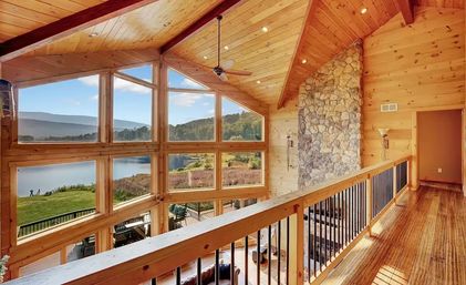 Sunlit lakefront mountain cabin loft with wood-paneled vaulted ceiling and stone fireplace, floor-to-ceiling windows framing lake and mountain views, hardwood floors and balcony railing.