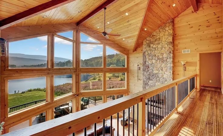 Sunlit lakefront mountain cabin loft with wood-paneled vaulted ceiling and stone fireplace, floor-to-ceiling windows framing lake and mountain views, hardwood floors and balcony railing.
