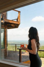 Person in a black dress with long wavy hair holding an amber cocktail, standing by a window overlooking a wooden deck, cabin balcony and sweeping mountain view under a blue sky.