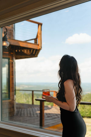 Person in a black dress with long wavy hair holding an amber cocktail, standing by a window overlooking a wooden deck, cabin balcony and sweeping mountain view under a blue sky.