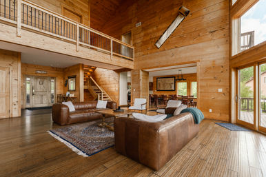 Cozy rustic log cabin living room with high wood-paneled walls, leather sofas and armchairs around a wooden coffee table on a patterned rug, loft balcony, open dining area and large windows opening to a deck and trees.