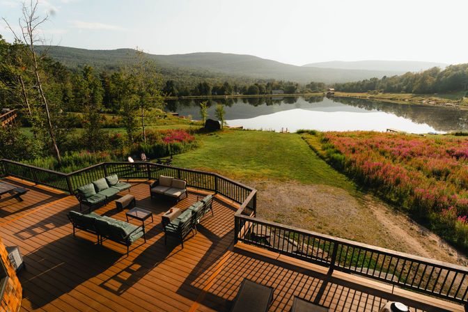 Cozy sunlit lakeside deck with outdoor patio seating overlooking a reflective mountain lake, grassy shore and pink wildflower meadow