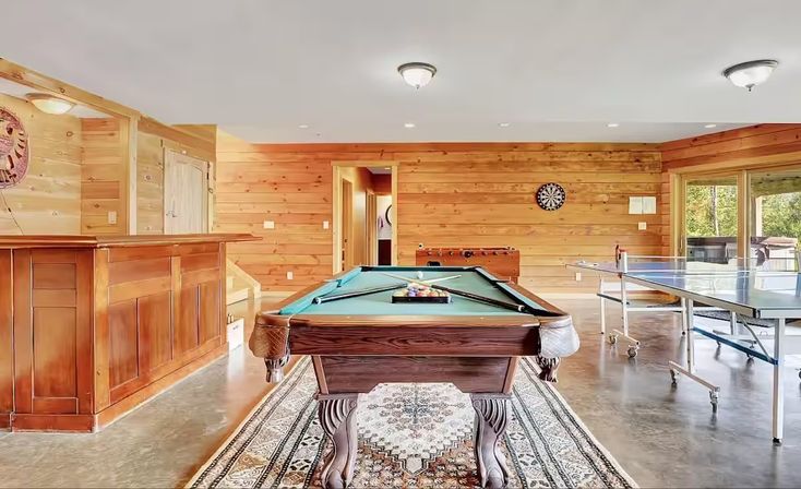 Cozy wood-paneled recreation room with a pool table front and center, bar on the left, ping-pong table, foosball, dartboard, rug, and large windows showing trees.