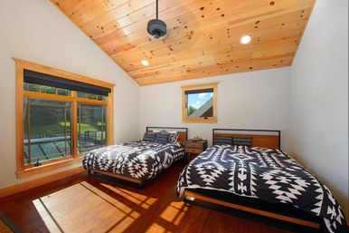 Cozy rustic bedroom with knotty pine vaulted ceiling, hardwood floors, two platform beds with black-and-white geometric bedding, a shared nightstand, and a large sunlit window overlooking a green yard.