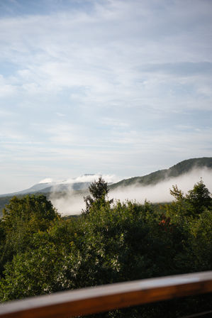 Scenic mountain landscape with green forested hills and a ribbon of morning fog drifting through the valley under a pale blue sky, viewed from a wooden balcony.