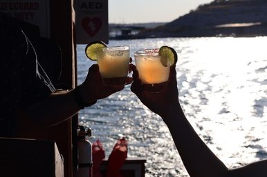 Two hands clinking lime-garnished cocktails at golden hour on a sunlit waterfront, silhouetted against sparkling water — a festive boat-side toast