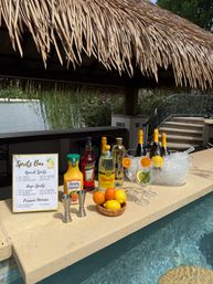 Sunny poolside tiki bar by a lake with a spritz station — Aperol and Prosecco bottles, orange juice, ice bucket, jiggers, and fresh citrus garnishes.