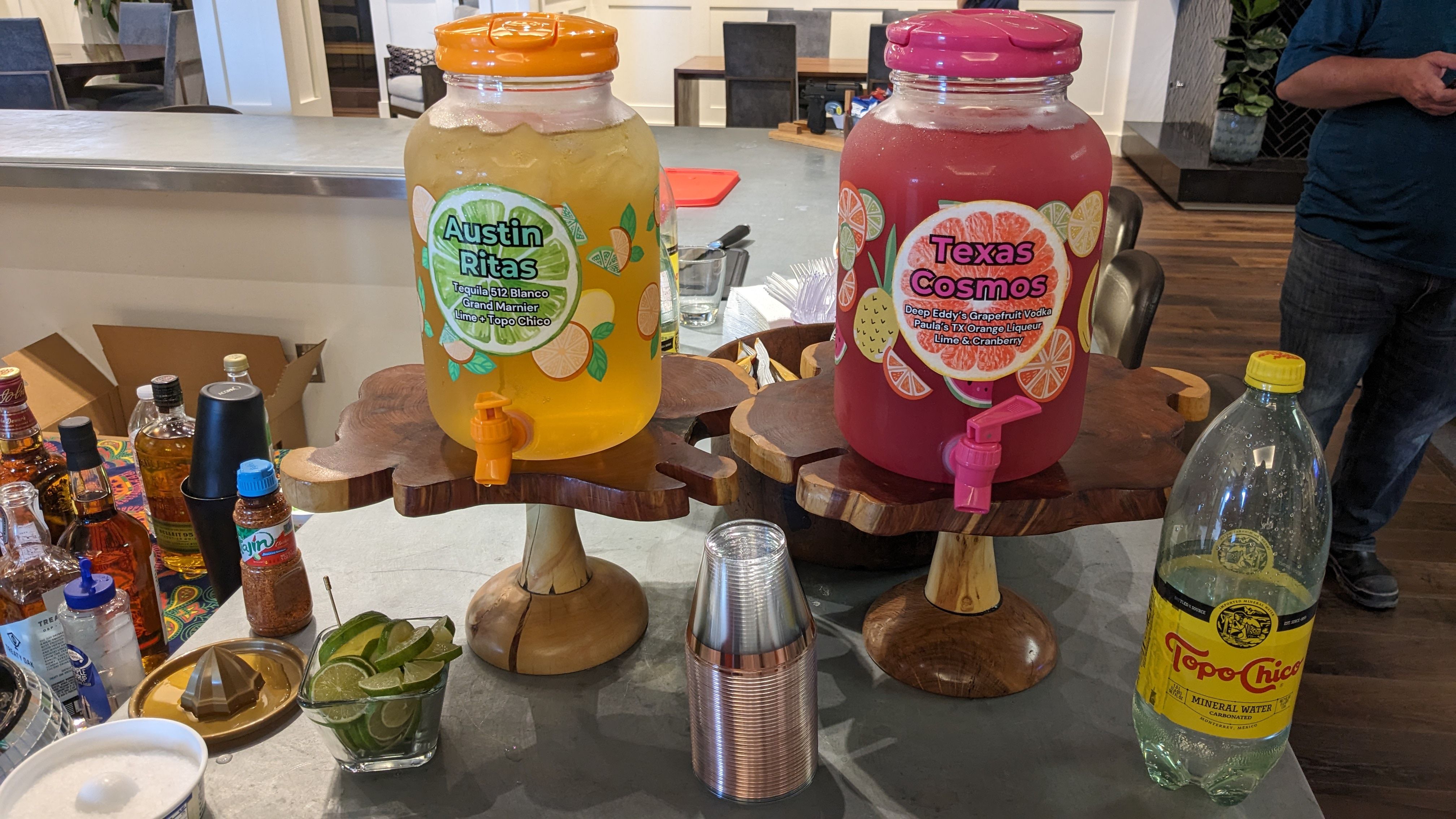 Two large glass drink dispensers (yellow and pink) on wooden stands at an indoor Texas cocktail station with lime wedges, stacked plastic cups and a mineral water bottle.
