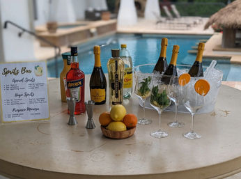 Poolside spritz bar setup at a tropical resort: bottles of aperitif and sparkling wine on a table, ice bucket with chilled bottles, wine glasses garnished with orange slices and mint, citrus bowl and recipe sign beside an outdoor swimming pool.
