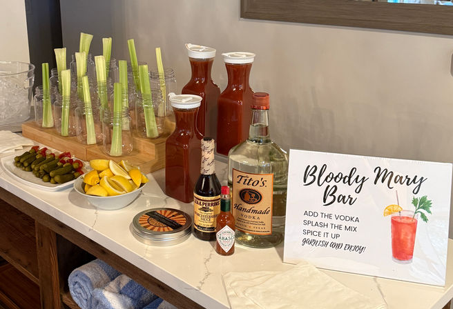 DIY Bloody Mary bar brunch setup on a white countertop with a vodka bottle, carafes of tomato mix, celery stalks in jars, lemon wedges, pickles, Worcestershire and hot sauce bottles, and a sign reading 'Bloody Mary Bar'.