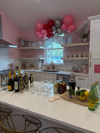 Pink modern home kitchen decorated for a celebration with metallic fringe and pink balloon garland above a white island stocked with bottles, glassware, citrus, and cocktail tools.