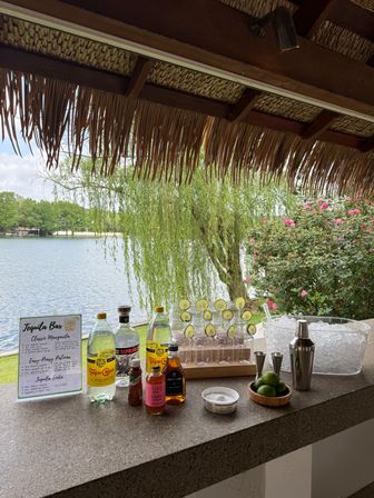 Lakeside tiki-style tequila bar setup on a stone counter with bottles, mixers, lime-garnished glasses, ice bucket and cocktail tools under a thatched roof, overlooking a willow tree and pink flowering bushes.