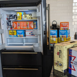 Open stainless-steel fridge in a modern kitchen with subway-tile backsplash, shelves packed with boxed seltzers and beer (Vizzy, Topo Chico, Miller Lite) and stacked cases on the counter next to a Keurig