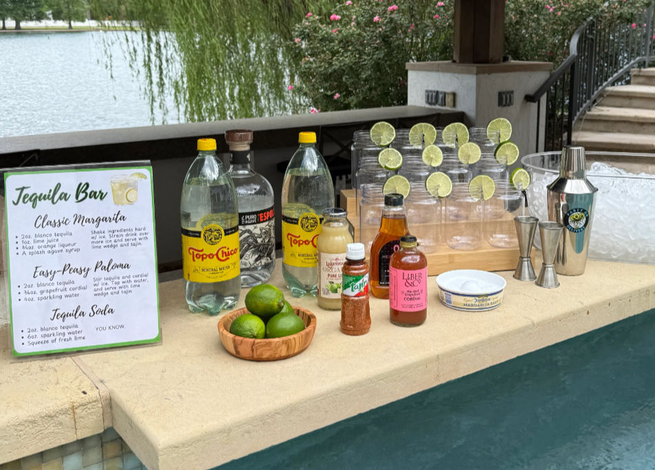 Poolside tequila bar setup on a lakeside patio with a margarita recipe sign, tequila and sparkling water bottles, a wooden bowl of limes, plastic glasses rimmed with lime wheels, cocktail shaker and jiggers.